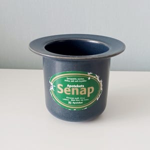 May include: A dark blue ceramic container with a wide rim and a label that reads "Apotekets Senap". The label is green and white with text in Swedish. The container is cylindrical and sits on a white surface.