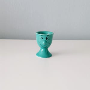May include: A teal ceramic egg cup with a whimsical face design. The cup features a winking eye, a red nose, and a smiling mouth. The egg cup is on a white surface.