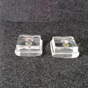 May include: Two clear, square glass candle holders. Each holder has a small circular indentation for a candle and a small sticker on the front. The holders are sitting on a dark gray surface.