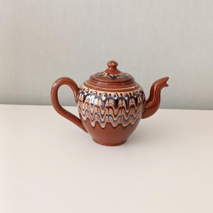Può includere: Una teiera in ceramica marrone con coperchio, manico e beccuccio. La teiera è decorata con un motivo ondulato nei toni del blu, bianco e marrone. La teiera è su una superficie bianca.