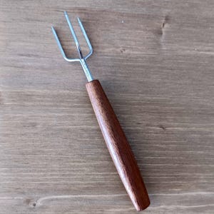 May include: A metal roasting fork with three tines and a wooden handle. The handle is a dark brown color and is smooth. The fork is designed for roasting marshmallows or other foods over a fire. The fork is on a wooden surface.