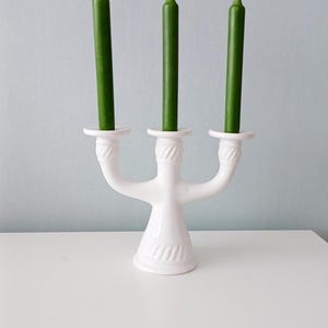 May include: A white ceramic candelabra with three arms, each holding a green taper candle. The candelabra has a decorative, textured design and sits on a white surface against a light blue background. The candles are approximately 15 cm tall.