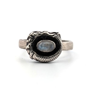 May include: A silver ring featuring a mermaid design. The ring has an oval-shaped, light blue gemstone set in a detailed bezel. The mermaid's face and tail are visible on the ring's setting, which is on a white background.