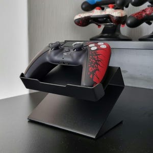 Könnte beinhalten: Ein schwarzer Metallständer, der einen schwarz-roten Videospiel-Controller hält. Der Controller hat ein rotes Design auf der rechten Seite. Weitere Controller sind im Hintergrund auf einem Regal zu sehen.