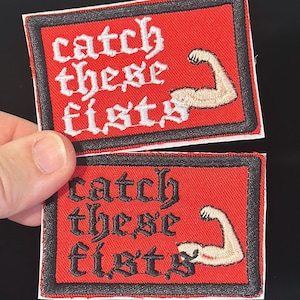 May include: Two red embroidered patches with the text "catch these fists" in white gothic lettering. Each patch features a flexed arm graphic. The patches have a black border.