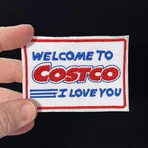 Puede incluir: Un parche rectangular blanco con un borde rojo y las palabras "WELCOME TO COSTCO I LOVE YOU" en azul y rojo. El logotipo de Costco se muestra prominentemente en rojo.