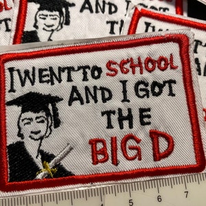 May include: A white embroidered patch with red stitching around the edges. The patch features a cartoon graduate holding a diploma and the text "I went to school and I got the big D".