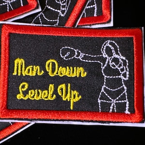 May include: Black patch with red border featuring the text "Man Down Level Up" in yellow script. A white outline of a boxer is on the right. Patches are a popular item.