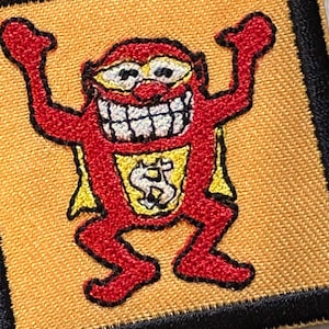 May include: Embroidered patch featuring a cartoon character in red with a yellow cape and a dollar sign on the chest. The character has a wide grin and is set against a gold background with a black border.