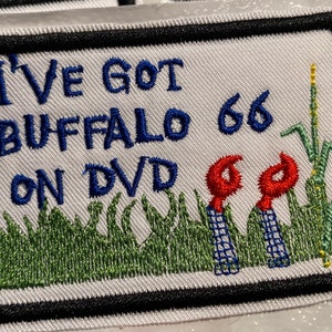 May include: Embroidered patch with the text "I'VE GOT BUFFALO 66 ON DVD" in blue thread. The patch features a black border, green grass, and red and blue stylized flowers. A yellow stalk of corn is also present.