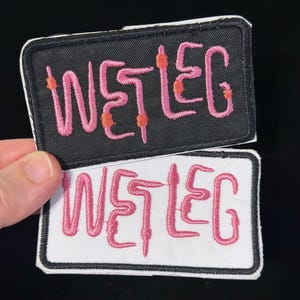 Puede incluir: Dos parches bordados con el texto "WET LEG" en rosa, letras estilizadas. Un parche tiene un fondo negro, el otro es blanco. Ambos parches tienen un borde blanco.