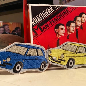 Może przedstawiać: Dwa haftowane naszywki z zabytkowymi samochodami. Niebieski samochód to hatchback, a żółty to sedan. Naszywki znajdują się na białym tle przed okładką albumu Kraftwerk.