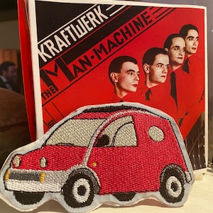May include: Red embroidered patch of a small car with a white roof and white wheels. The patch is on a white background. The car is similar to a Volkswagen.