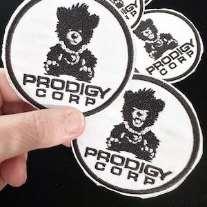 May include: A collection of white, circular patches with a black and white bear illustration and the text "PRODIGY CORP." The bear has a necklace and is centered within a black border.