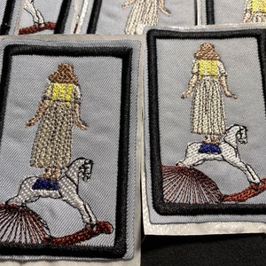 May include: A set of embroidered patches featuring a person standing on a rocking horse. The patches are on a light blue background with black stitching around the edges. The person is wearing a white dress with a yellow vest and has brown hair. The rocking horse is white with a blue saddle.