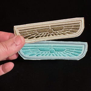 May include: Two embroidered patches with a winged design. One patch is tan and brown, the other is teal and light blue. The patches have a curved rectangular shape with a detailed, symmetrical wing pattern. The patches are likely for clothing or accessories.