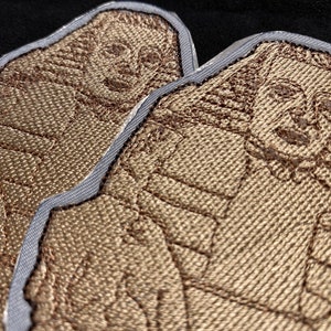 May include: Two brown embroidered patches featuring a detailed design of a person's face. The patches have a light grey border.
