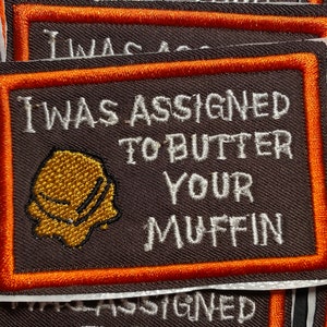 Puede incluir: Un parche bordado marrón y naranja con el texto "I was assigned to butter your muffin". El parche presenta un pequeño muffin amarillo de estilo de dibujos animados.