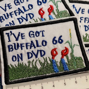 May include: A white embroidered patch with black trim, featuring the text "I've Got Buffalo 66 On DVD" in blue and red lettering. The patch depicts two red buffalo heads with blue bodies, standing in a field of green grass.