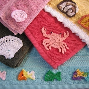 Op de afbeelding: Een set van vijf gehaakte handdoeken met een zeethema. De handdoeken zijn in verschillende kleuren, waaronder roze, rood, geel en blauw. De gehaakte ontwerpen omvatten een schelp, een krab en een vis.