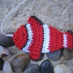May include: A crocheted red, white, and brown fish with a white string attached to the top. The fish is lying on a bed of sand with several smooth, gray rocks.