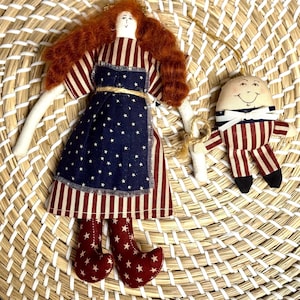Könnte beinhalten: Zwei handgefertigte patriotische Ornamente. Eines ist eine Puppe mit rotem Haar, einem gestreiften Kleid und einer blauen, sternenbesetzten Schürze. Das andere ist eine eiförmige Figur mit einem lächelnden Gesicht, die ein gestreiftes Outfit trägt. Beide befinden sich auf einem geflochtenen Korb.