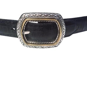 May include: A black belt with a textured crocodile-embossed finish featuring a rectangular buckle. The buckle has a silver-toned outer frame with a woven pattern and a smooth gold-toned inner border surrounding the central prong mechanism.