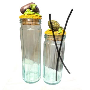 Könnte beinhalten: Zwei hohe, sechseckige Vorratsgläser aus klarem Glas mit Korkdeckeln. Das größere Glas hat einen geformten Auberginen-Aufsatz, das kleinere einen Artischocken-Aufsatz. Beide Deckel haben eine texturierte, gelbe, sonnenblumenartige Basis.
