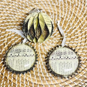 Pode incluir: Três ornamentos redondos com partituras vintage e texto impresso em papel creme. Um ornamento é uma esfera 3D com bordas de glitter preto, enquanto dois são discos circulares planos com bordas recortadas de glitter preto e fitas brancas.