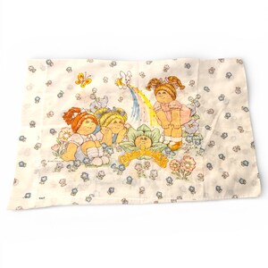May include: A rectangular, off-white pillowcase featuring a colorful illustration of Cabbage Patch Kids. The design includes three dolls, a rainbow, flowers, and butterflies. The words "Cabbage Patch Kids" are also visible.