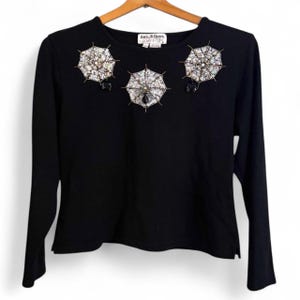 May include: Black long-sleeve shirt with three embellished spiderweb designs on the front. Each spiderweb is intricately detailed with sparkling beads and crystals. The shirt has a classic crew neck and a straight hemline.