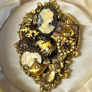 Puede incluir: Un broche dorado ornamentado que presenta un camafeo central en blanco y negro de un perfil, un cabujón ovalado negro con una ilustración de ciervo dorado, una esfera de reloj pequeña, un dije de bolsa de dinero, una lámpara, flores y brillantes rojos.