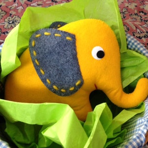 May include: A yellow felt elephant toy with a gray felt ear and a white eye, sitting in a blue and white gingham basket lined with green tissue paper.