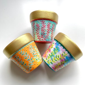 May include: Three small terracotta pots with gold rims. Each pot is painted with a different design featuring green leaves and a colourful background. The pots are on a white surface.