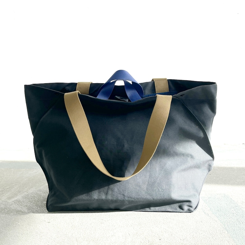 May include: A large, dark grey canvas tote bag with tan straps and a blue accent on the top. The bag is sitting on a white surface.