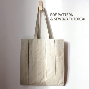 Puede incluir: Un bolso tote beige con pliegues verticales cuelga de una clavija de madera. El bolso tiene forma rectangular con un asa. El texto a la derecha dice "PDF PATTERN & SEWING TUTORIAL".