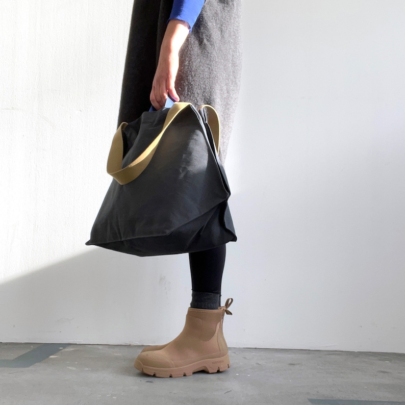 May include: A person is holding a large, dark grey tote bag with a light brown strap. The bag is made of a canvas-like material and has a slightly slouchy shape. The person is wearing brown ankle boots.