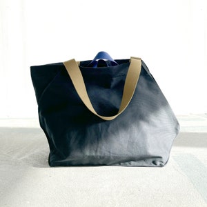 May include: A large, dark grey canvas tote bag with two tan straps. The bag is empty and sitting on a white surface.
