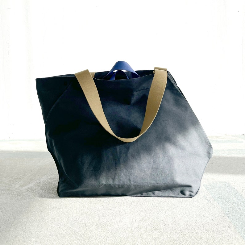 May include: A large, dark grey canvas tote bag with two tan straps. The bag is empty and sitting on a white surface.