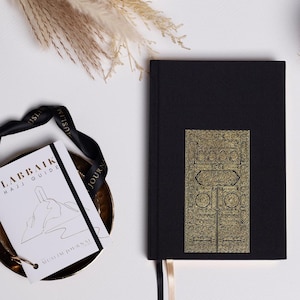 May include: A black journal with a gold embossed design of the Kaaba, a cube-shaped building in Mecca, Saudi Arabia. The journal has the text "Labbaik Hajj Guide" and "Muslim Journal" on the cover.