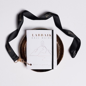 May include: A white booklet with the title "Labbaik Hajj Guide" and a black and white illustration of a mountain with a building on top. The booklet is on a gold-coloured plate and is tied with a black ribbon with the text "The Muslim Journal" printed in gold.