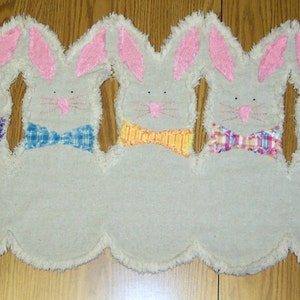 Op de afbeelding: Een witte stoffen konijnendeurstopper met een gekrulde rand. Het konijn heeft roze oren en een strik in blauw, paars, geel of ruit.