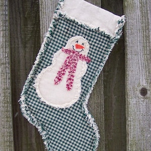 Peut inclure: Une chaussette de Noël faite à la main avec un motif à carreaux verts et blancs. Elle présente un appliqué de bonhomme de neige blanc avec une écharpe rose et un nez orange. Les bords sont effilochés, lui donnant un aspect rustique. Une boucle est attachée pour l'accrocher.