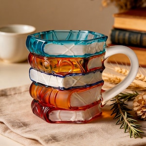 Pode incluir: Uma caneca de café única em forma de pilha de livros coloridos. A caneca tem uma alça branca e é feita de vidro transparente em tons de azul, laranja e vermelho. O design inclui detalhes como lombadas e páginas de livros.