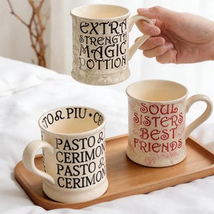May include: Three cream-colored ceramic mugs with curved handles. One mug reads "EXTRA STRENGTH MAGIC POTION" in black. Another says "SOUL SISTERS BEST FRIENDS" in red. The third mug has text in black. All are on a wooden tray.