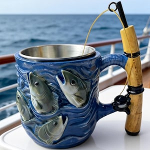 Pode incluir: Caneca de cerâmica azul com interior em aço inoxidável, com um design em relevo de peixes nadando em ondas. A alça é uma vara de pesca, completa com carretel e linha. A caneca está em uma superfície branca com um fundo de oceano azul.