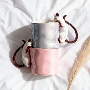 Puede incluir: Dos tazas de cer&aacute;mica apiladas. La superior es gris y blanca, la inferior rosa. Cada taza tiene un gato blanco trepando por el lateral, formando un asa marr&oacute;n. Dise&ntilde;o original y decorativo.