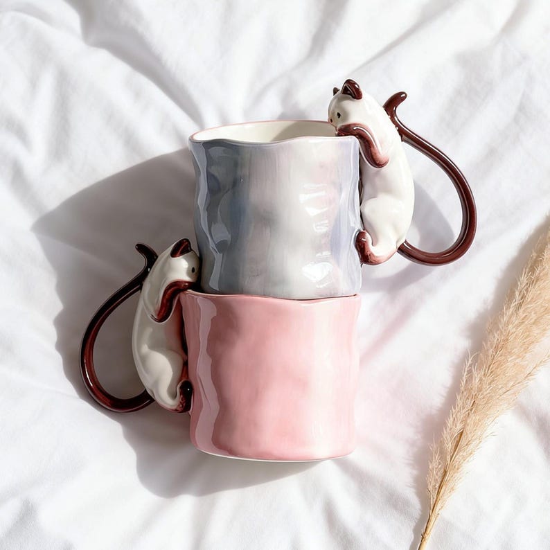 Puede incluir: Dos tazas de cer&aacute;mica apiladas. La superior es gris y blanca, la inferior rosa. Cada taza tiene un gato blanco trepando por el lateral, formando un asa marr&oacute;n. Dise&ntilde;o original y decorativo.