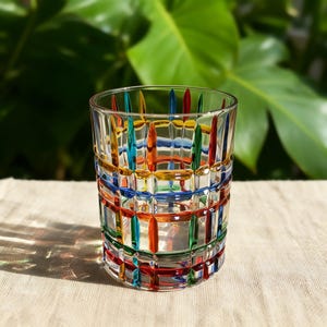 May include: A clear, patterned drinking glass with a colorful grid design. The glass features vertical and horizontal lines in red, orange, yellow, green, blue, and teal. The glass sits on a neutral-colored surface with a blurred green background.
