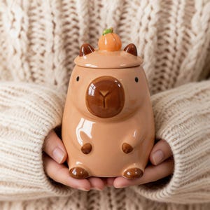 May include: A ceramic capybara-shaped mug with a lid, featuring a pumpkin on top. The mug is light brown with darker brown accents and small black eyes. The mug is held in front of a cream-colored sweater.